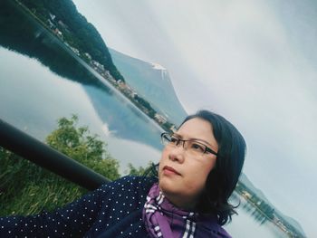 Tilt image of thoughtful mature woman looking away while standing by lake against sky