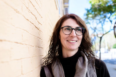 Smiling woman with eyeglasses leaning on wall, looking away