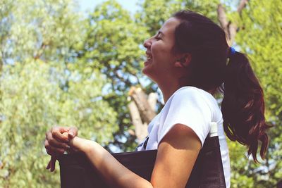 Side view of woman laughing while sitting on chair against trees