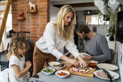 Girl looking at mother preparing pizza standing at table