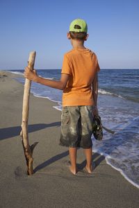 Rear view of boy holding stick and standing on shore against clear sky