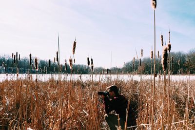 Side view of young man photographing while kneeling at field against sky