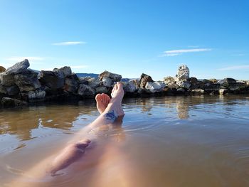 Low section of woman sitting on rock by sea against blue sky