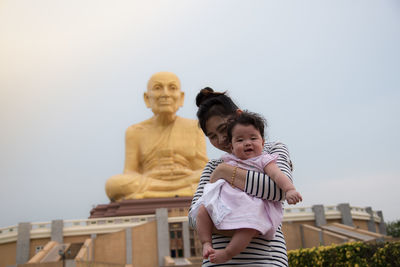 Smiling mother embracing cute daughter while standing against statue