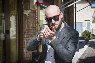 Young man with a shaved head, beard, and sunglasses, lights up a cigar