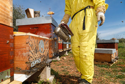 Cropped unrecognizable beekeepers in protective costume and mask using smoker while inspecting honeycomb in apiary  person