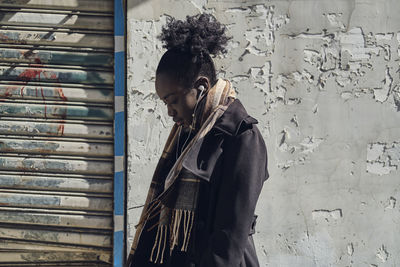 Side view of young attentive ethnic female in scarf with afro hair bun listening to song in earphones in town while looking down