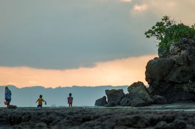 People on rock by sea against sky during sunset