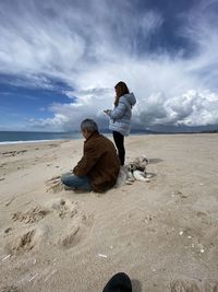 Rear view of friends sitting on sand at beach against sky