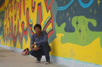 Full length portrait of young man crouching by graffiti wall