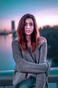 Portrait of beautiful young woman standing against river in city during sunset