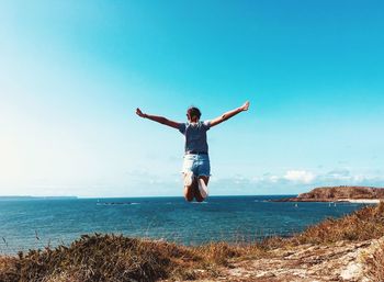 Rear view of woman jumping on mountain by sea against blue sky