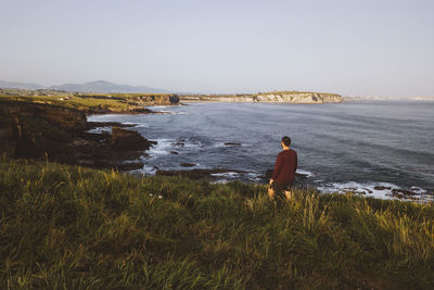 A young man stands in front of a cliff starting at the coastline, cantabria, spain