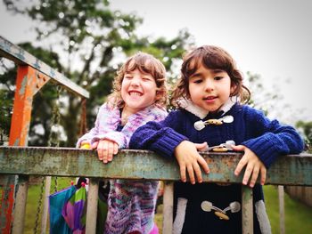 Portrait of happy girls standing by railing at park against sky