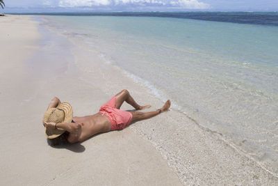 Fit man with sun hat, tanning on sandy beach with clear blue waters