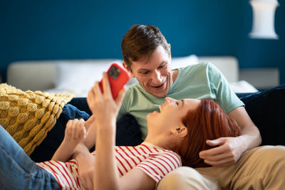 German man and woman with smartphone laughing at funny videos internet lying on sofa.