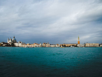 Cityscape by sea against cloudy sky