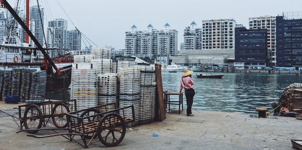 Rear view of man standing by crates against lake in city