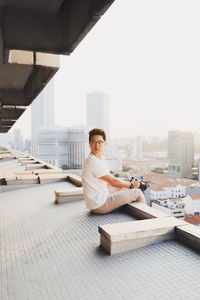 Side view of young man holding digital camera while sitting on floor in city