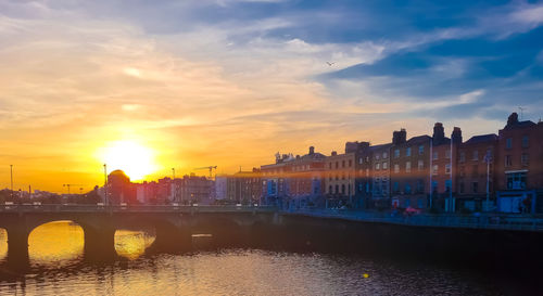 Bridge over river by buildings against sky during sunset