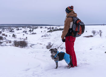 Man with backpack with mixed breed dog in warm blue suit walking in snow mountain in winter