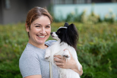 Caucasian red-haired woman holding pappilion dog in her arms outdoors. black and white continental