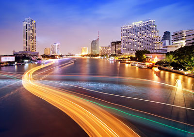 Light trails on road by buildings against sky at night