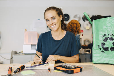 Portrait of smiling female technician working while sitting at workbench in creative office