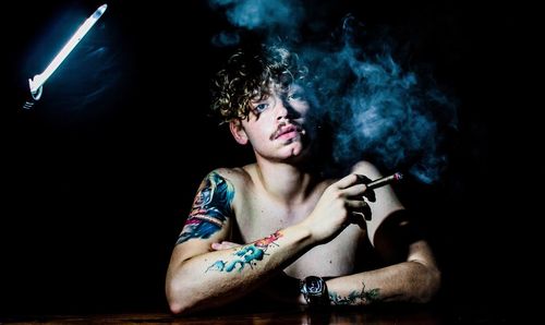 Portrait of shirtless tattooed man smoking cigar at table against black background