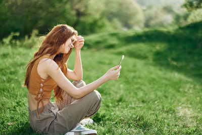 Side view of young woman using mobile phone while sitting on grassy field