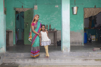 Full length of a smiling mother and daughter standing in front of house doorway in a rural  india