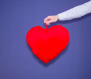 Close-up of hand holding red heart shape pillow against purple wall
