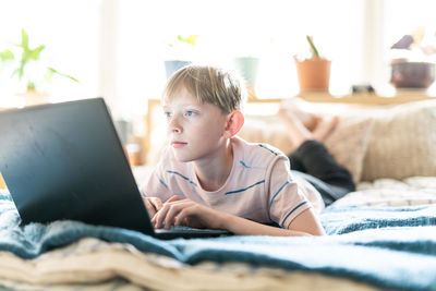 Teen boy working on laptop while lying on bed in brightly lit space