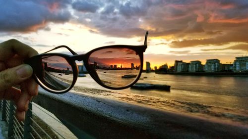 Reflection of hand holding sunglasses against buildings in city during sunset