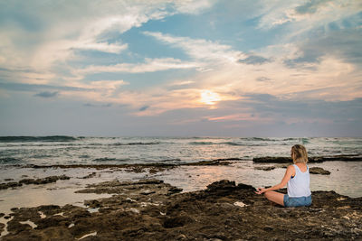 Woman doing yoga at beach against sky during sunset