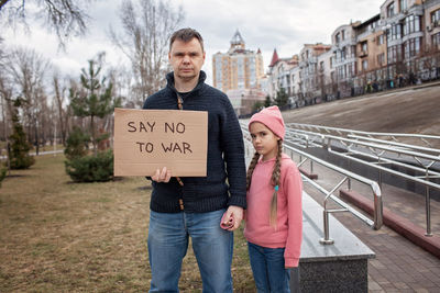 Middle aged father with daughter holding a poster with anti-war message over cityscape background