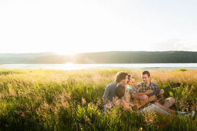 Friends listening to man playing guitar while sitting amidst grass