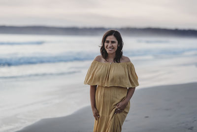 Lifestyle portrait of young woman walking on beach against cloudy sky