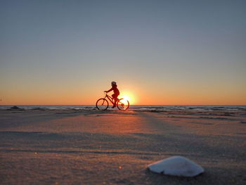 Girl riding bicycle at beach against sky during sunset