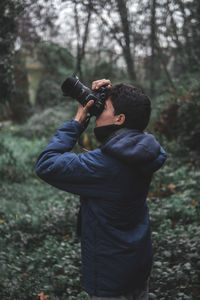 Side view of young man photographing while standing in forest