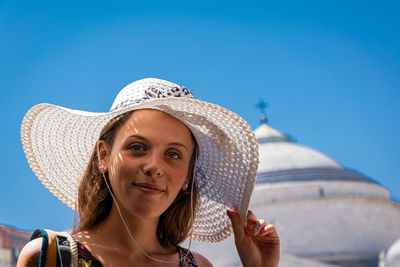 Low angle portrait of young woman wearing hat against clear blue sky