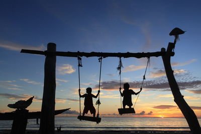 Silhouette of boys on swing by sea against sky during sunset