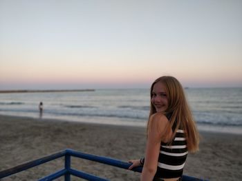 Portrait of teenage girl standing by railing at beach during sunset