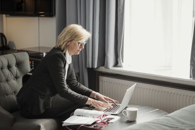 Side view of businesswoman using laptop by window at hotel room