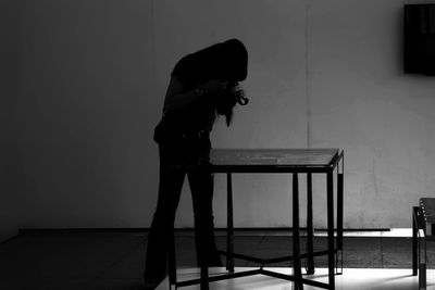 Woman photographing while standing by table against wall