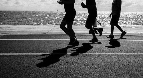 Low section of silhouette people on footpath by sea during sunny day