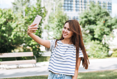 Portrait of young woman student with long hair taking selfie with mobile phone in city park