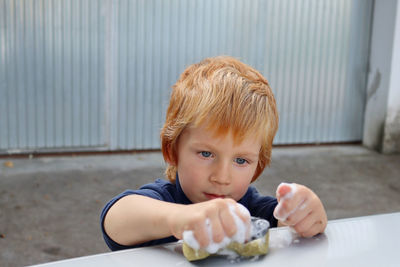 The blond boy stares intently at the soapy sponge. a  boy squeezes a sponge tightly in his hand.