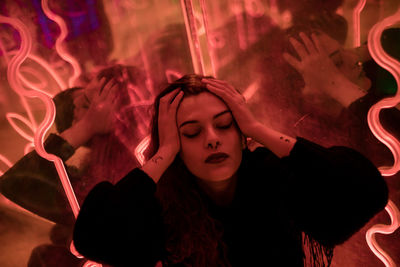 Stylish long haired melancholic brunette in casual wear closed eyes among neon signs at city street