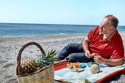 Middle aged couple having a picnic at the seaside with fresh exotic fruit. man cuts a melon.